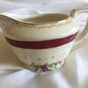 May include: Vintage cream-coloured ceramic creamer with a burgundy stripe and gold trim. It features a handle, spout, and floral base accents. This creamer is suitable for serving cream or milk, and is likely vintage.