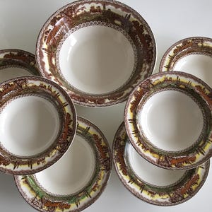 May include: A set of six white ceramic bowls with a brown and yellow landscape design around the rims. The bowls are arranged on a white surface, with one larger bowl in the center and five smaller bowls surrounding it.