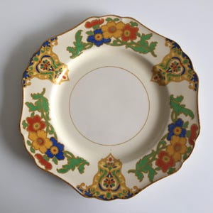 May include: A cream-colored ceramic plate with a scalloped edge, featuring a decorative border of vibrant red, yellow, and blue flowers intertwined with green foliage and gold-toned accents. The center of the plate is plain and circular.