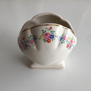 May include: A cream-colored ceramic bowl with a scalloped rim and a gold-trimmed edge. The exterior features a decorative floral pattern consisting of pink roses, blue cornflowers, and small yellow and red blossoms arranged in a horizontal band.