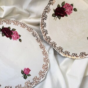 May include: Two off-white plates with a floral rose design. The plates have a scalloped edge with a gold and brown floral border. Each plate features a cluster of roses in shades of pink and burgundy.