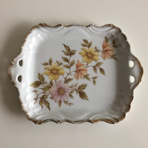 May include: A white porcelain serving tray with a scalloped edge and gold trim. The tray features two side handles and a floral illustration of yellow, peach, and pink flowers with green leaves arranged diagonally across the center surface.