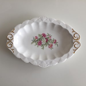 以下が含まれることがあります： 白の楕円形サービング皿、スカラップエッジとゴールドのアクセント。 皿の中央には、ピンク、白、緑の花のブーケがあります。 食べ物を提供したり、アイテムを展示したりするための装飾品です。