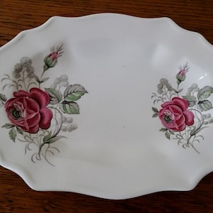 May include: White ceramic serving platter with a scalloped edge. The platter features a floral design with red roses, green leaves, and grey accents. The roses are the focal point, adding a touch of elegance to the piece.