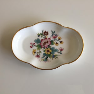 May include: A white ceramic dish with a gold rim and an oval shape. The dish is decorated with a floral pattern in pink, yellow, and green. The flowers are arranged in a central cluster, providing a decorative element.