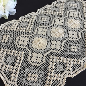 Op de afbeelding: Crèmekleurige kanten tafelloper met een gedetailleerd bloemen- en geometrisch patroon. De loper heeft geborduurde bloemen en een delicaat, open ontwerp. De randen zijn geschulpt, wat een decoratief tintje geeft aan dit textiel in vintage stijl.