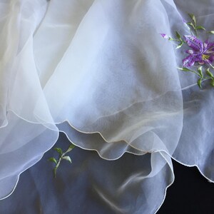 以下が含まれることがあります： 白いシアーなスカーフ。縁はスカラップで、紫の花と緑の葉の刺繍が施されています。軽くて流れるような生地で、装飾的なアクセサリーに適しています。生地は透明です。