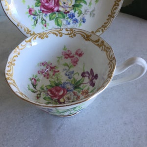 Puede incluir: Juego de taza y platillo blancos con un diseño floral de rosas rosas, rojas, azules y amarillas. La taza tiene un borde y asa dorados. El platillo también es blanco con un diseño floral a juego.