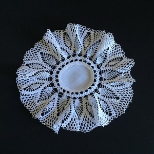 Puede incluir: Un tapete blanco circular con un intrincado diseño de encaje. El tapete presenta un círculo central blanco sólido rodeado por un patrón calado detallado. Los bordes están festoneados, agregando un toque delicado. El tapete está sobre un fondo negro.