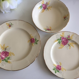 May include: A set of three vintage ceramic plates featuring a floral design with pink roses and yellow mimosa flowers. The plates have a cream-coloured background and a gold rim.