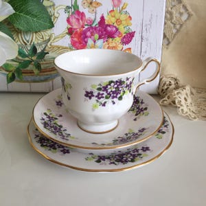 以下が含まれることがあります： 紫と緑の花柄が施された、白いティーカップ、ソーサー、サイドプレートのセット。縁は金でトリミングされています。ティーカップはカーブしたハンドルと台座が付いています。背景には花の絵が描かれています。