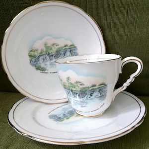 May include: A white teacup and saucer set with a gold trim. The cup and saucer feature a watercolor image of a waterfall with the text "CASCADE MILLSOW", "NEW RIVER GORGE,  W.VA." and "WEST VIRGINIA STATE PARKS"