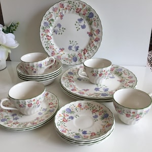 May include: A set of white ceramic tableware featuring a floral design. The collection includes plates, saucers, and teacups, decorated with pink, purple, and blue flowers. Each piece has a green border around the rim, offering a traditional look.