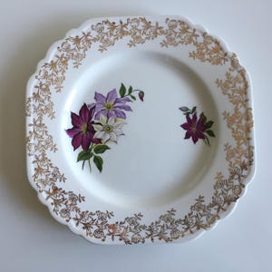 May include: White square plate with a scalloped edge and gold floral trim. The plate features a cluster of purple, white, and green flowers in the centre and a single purple flower on the right. A decorative serving plate.