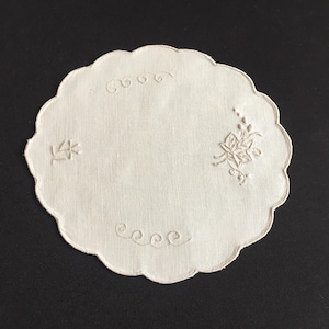 Pode incluir: Base para copos branco sujo com borda ondulada e desenhos bordados de flores e rolos. A base para copos é redonda e feita de um tecido macio. O bordado é em bege claro, adicionando um toque delicado.