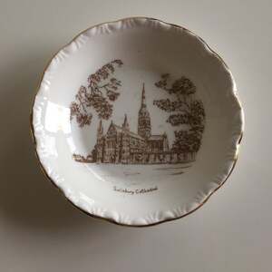 May include: A white ceramic dish with a gold rim features a brown line drawing of Salisbury Cathedral. The dish is inscribed with the text "Salisbury Cathedral".