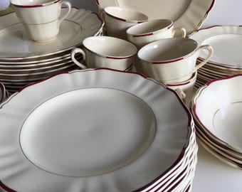 May include: A collection of cream-colored dinnerware with a scalloped edge and a burgundy trim. The set includes plates, bowls, and teacups, arranged in stacks and groupings. The teacups have a matching burgundy trim and a gold accent line.