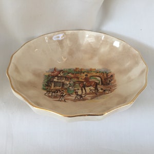 May include: Cream-colored, scalloped-edge dish with a gold rim. The center features a detailed illustration of a hunting scene with people, dogs, a horse, and a bridge. A decorative piece.