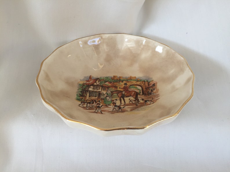 May include: Cream-colored, scalloped-edge dish with a gold rim. The center features a detailed illustration of a hunting scene with people, dogs, a horse, and a bridge. A decorative piece.
