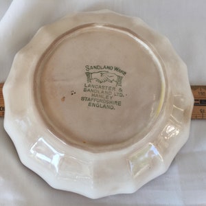 May include: A vintage, off-white Sandland Ware plate with a scalloped edge. The plate features the text "SANDLAND WARE" and "LANCASTER & SANDLAND LTD. HANLEY STAFFORDSHIRE ENGLAND." A wooden ruler is next to the plate.