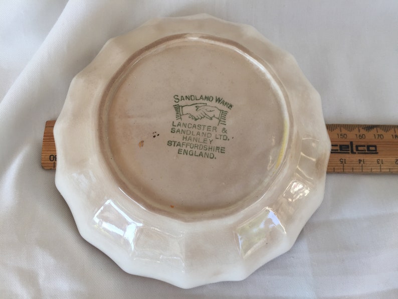 May include: A vintage, off-white Sandland Ware plate with a scalloped edge. The plate features the text "SANDLAND WARE" and "LANCASTER & SANDLAND LTD. HANLEY STAFFORDSHIRE ENGLAND." A wooden ruler is next to the plate.