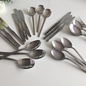 Puede incluir: Un surtido de cubiertos antiguos, que incluye cucharas, cuchillos y tenedores, dispuestos sobre una superficie blanca. La cubertería tiene un acabado metálico brillante, con algunas piezas que presentan patrones florales decorativos en los mangos. Se ven varios tamaños de cuchara.