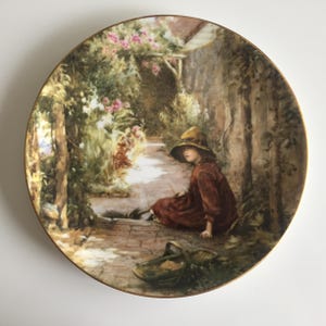 May include: A round decorative ceramic plate with a gold-trimmed edge featuring a detailed illustration of a child wearing a wide-brimmed hat and a dark red dress, sitting on a brick path in a garden with pink flowers and a woven basket nearby.