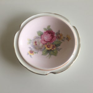 May include: A small, white ceramic dish with a pink interior and a gold rim. The dish features a floral design with pink, white, and yellow roses.
