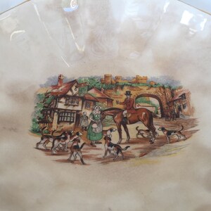May include: A cream-colored, scalloped-edge decorative bowl with a detailed illustration. The illustration depicts a hunting scene with a rider on horseback, hounds, and figures in period clothing near a village.