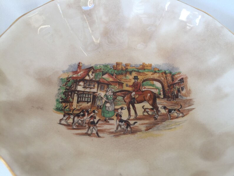 May include: A cream-colored, scalloped-edge decorative bowl with a detailed illustration. The illustration depicts a hunting scene with a rider on horseback, hounds, and figures in period clothing near a village.