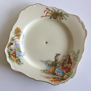 May include: Cream-colored, scalloped-edge plate with a gold trim. The plate features a painted scene of a couple in traditional Scottish attire, a landscape, and a bagpipe illustration. A small hole is in the center.