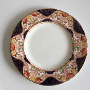 May include: A round ceramic plate featuring a plain white center surrounded by a decorative border. The border displays a pattern of red and yellow roses, green foliage, and dark blue sections with a fine gold crackle texture and gold trim.