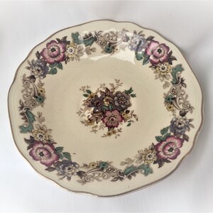 Puede incluir: Plato crema con borde festoneado y diseño floral. El plato presenta un arreglo floral central y un borde de flores rosas, moradas y amarillas con detalles verdes. El borde tiene un ribete dorado.