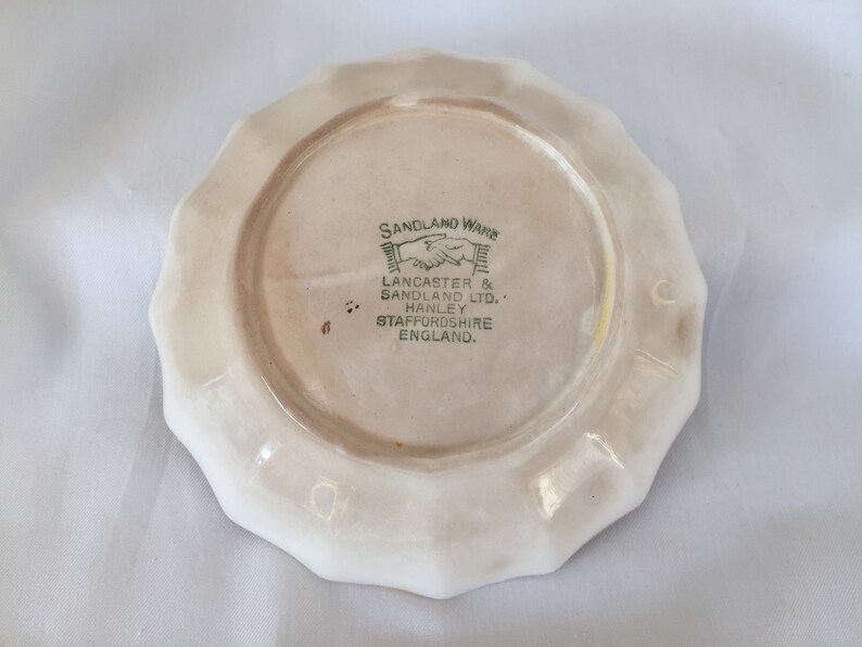 May include: A cream-colored, scalloped-edge plate with the text "SANDLAND WARE" and "LANCASTER & SANDLAND LTD. HANLEY STAFFORDSHIRE ENGLAND" in green. The plate has a light brown center.