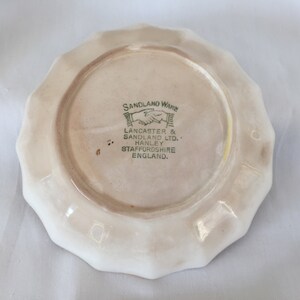 May include: A small, off-white ceramic plate with a scalloped edge. The underside features a central circular area with text in green and black, including "SANDLAND WARE" and "LANCASTER & SANDLAND LTD."