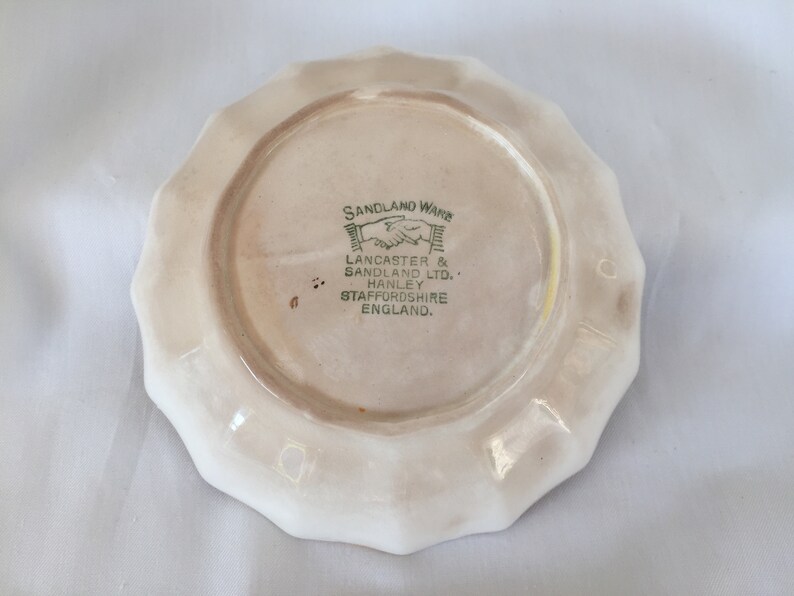 May include: A small, off-white ceramic plate with a scalloped edge. The underside features a central circular area with text in green and black, including "SANDLAND WARE" and "LANCASTER & SANDLAND LTD."