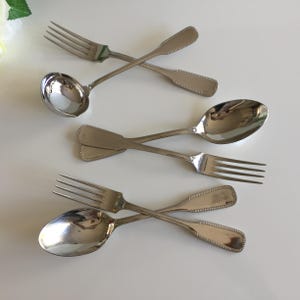 May include: A collection of vintage silverware, including forks, spoons, and a ladle, arranged on a white surface. The silverware has a shiny, reflective surface and a decorative beaded edge. The utensils are crossed over each other.
