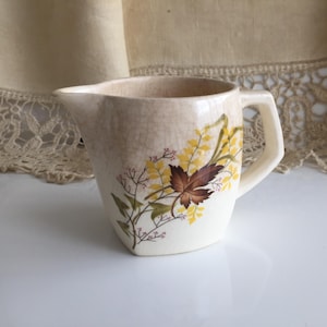 Könnte beinhalten: Cremefarbener Keramikkrug mit floralem Design. Der Krug hat ein braunes Blatt und gelbe Blüten mit grünen Stielen. Der Griff ist eckig und der Krug hat einen Ausguss zum Ausgießen. Die Oberfläche hat eine Craquelé-Glasur.