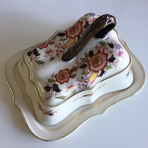 以下が含まれることがあります： 赤、オレンジ、青の花柄が施された白いセラミックバターディッシュ。 茶色の取っ手が付いた蓋があり、お揃いのスカラップトレイの上に置かれています。縁は金でトリミングされています。