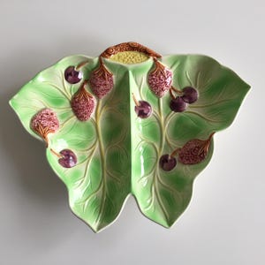 Könnte beinhalten: Dekorative Keramik-Servierplatte in Blattform, mit hellgrüner Basis und erhabenen Details. Die Platte ist mit rosa Erdbeeren und dunkelvioletten Kirschen verziert. Oben befindet sich ein brauner Griff.