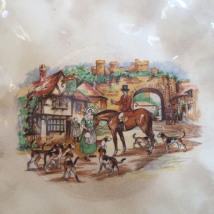 May include: A decorative plate with a detailed illustration of a hunting scene. The image depicts a man on horseback, hounds, and figures in a village setting with a castle in the background. The plate has a cream-colored background.