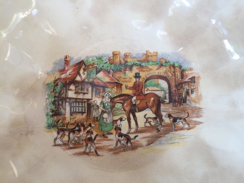 May include: A decorative plate with a detailed illustration of a hunting scene. The image depicts a man on horseback, hounds, and figures in a village setting with a castle in the background. The plate has a cream-colored background.