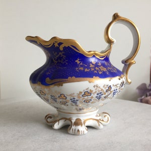 Könnte beinhalten: Eine Porzellan-Sahnekännchen mit blau-weißem Korpus, verziert mit goldenen Blumenmustern und Verzierungen. Das Kännchen hat einen gebogenen Griff und einen dekorativen Sockel, der ein elegantes Design zeigt.
