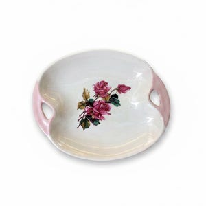 May include: An oval ceramic serving dish with a white center and soft pink handles on each side. The center of the dish features a detailed illustration of pink roses with green leaves and stems, rendered in a realistic style on the glossy surface.