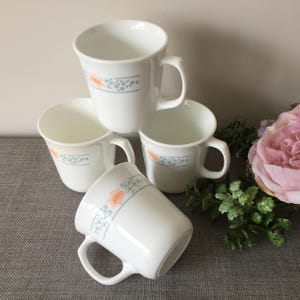 May include: A set of four white ceramic mugs featuring a delicate floral pattern with light blue leaves and soft orange blossoms. Each mug has a smooth, tapered body and a curved handle, arranged in a stacked formation to display the consistent design.