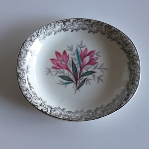 May include: A vintage, off-white ceramic dish with a floral design. The center features a cluster of pink lilies with green and blue accents. The rim is decorated with a silver and black ornate pattern. A classic piece for serving or display.