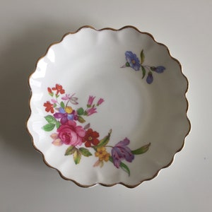 May include: A white porcelain trinket dish with a scalloped edge and a gold trim. The dish features a floral design with pink, red, purple, and blue flowers.