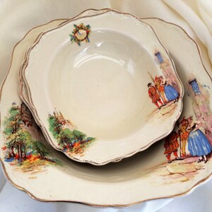 May include: Two off-white, scalloped-edge serving dishes. The larger dish has a smaller bowl nested inside. Both have a gold trim and feature a colourful, vintage illustration of a European village scene with people in period clothing.