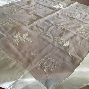 Può includere: Una tovaglia quadrata con un pannello centrale trasparente color beige chiaro decorato con motivi a foglia bianchi ricamati. Il tessuto è incorniciato da un bordo opaco color beige chiaro simile al raso che crea un contrasto di consistenza.