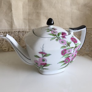 Könnte beinhalten: Eine weiße Porzellan-Teekanne mit einem Blumenmuster aus rosa und magentafarbenen Blüten und grünen Blättern. Die Teekanne hat einen schwarzen Griff, einen schwarzen Knauf auf dem Deckel und einen schwarz abgesetzten Ausguss. Der Körper ist facettiert.